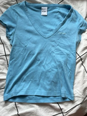 Blå v-ringad topp från Iets Frans - Snygg ljusblå topp från Iets Frans med v-ringning och korta ärmar. Tillverkad i mjuk bomull som känns skön mot huden. Enkel och stilren design med liten broderad logga på bröstet. Perfekt att matcha med jeans eller kjol.