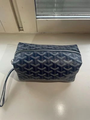 Blå necessär från G*oyard - Helt ny Snygg blå necessär från G*oyard med det klassiska mönstret i vitt och grått. Väskan har en rektangulär form, dragkedja upptill och en praktisk handledsrem. Perfekt för att organisera småsaker eller smink. Tillverkad i tåligt canvasmaterial.