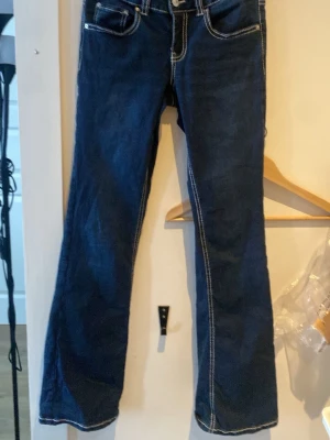 Mörkblå bootcut jeans med kontrastsömmar - Säljer ett par mörkblå bootcut jeans med vita kontrastsömmar och snygga fickdetaljer bak. Klassisk femficksmodell i denim med knapp och dragkedja framtill. Perfekta för en avslappnad och trendig look.