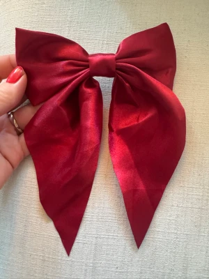 Stor vinröd hårrosett i satin - Snygg och stor hårrosett i vinröd satin med metallklämma på baksidan. Perfekt accessoar för att piffa upp frisyren och ge en elegant touch. Rosetten har en klassisk form med långa band och glänsande finish. Har bundlerabatt vid köp av fler varor🥰