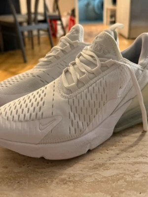 Vita Nike Air Max 270 sneakers - Säljer ett par fräscha vita Nike Air Max 270 sneakers med ventilerande mesh-ovandel, synlig Air-enhet i hälen och klassisk Nike-logga på sidan. Skorna har en sportig look med vita snören och detaljer i plast. Perfekta för dig som gillar stilrena och bekväma sneakers.