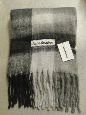 Acne Studios halsduk - Säljer en fluffig halsduk från Acne Studios i rutigt mönster med nyanser av svart, grått och vitt. Halsduken har långa fransar i ändarna och en tydlig Acne Studios-logga. Perfekt accessoar för att lyfta din outfit.