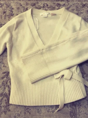 Beige omlottstickad tröja med knytband - Snygg beige stickad omlott-tröja med djup V-ringning och bred ribbad mudd i nederkant och ärmslut. Tröjan har ett knytband i midjan som ger en trendig och feminin look. Storlek XS från Gina tricot 