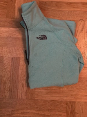 Ljusblå fleece från The North Face - Mysig ljusblå fleecejacka från The North Face med halv dragkedja och hög krage. Perfekt för kyliga dagar och enkel att slänga på sig. Tillverkad i mjukt fleecematerial och har en diskret logga på bröstet.