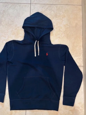 Mörkblå hoodie från Polo Ralph Lauren - Snygg mörkblå hoodie från Polo Ralph Lauren med klassisk orange broderad logga på bröstet. Hoodien har huva med dragsko och är tillverkad i mjukt bomullsmaterial som känns riktigt skönt mot huden. Perfekt för dig som gillar stilrena och tidlösa plagg.