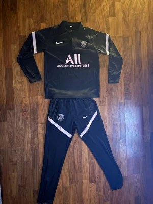 Svart PSG träningsset från Nike - Säljer ett svart PSG träningsset från Nike med långärmad tröja och matchande byxor. Setet har vita och rosa detaljer, PSG-logga och Nike Swoosh på både tröja och byxor. Materialet är syntetiskt och snabbtorkande, perfekt för träning.