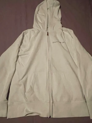 Beige hoodie med dragkedja från Gant - Snygg beige hoodie från Gant med dragkedja framtill och broderad text 'GANT 1949' på bröstet. Hoodien har två fickor fram och en klassisk huva. Perfekt för en chill och avslappnad stil.