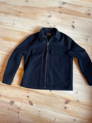 Svart overshirt från H&M med dragkedja - Stilren svart overshirt från H&M med två stora bröstfickor och hel dragkedja framtill. Overshirten har klassisk krage och är tillverkad i ett tjockare tyg med mjuk känsla, perfekt för lager på lager. Passar dig som gillar enkel och clean stil.