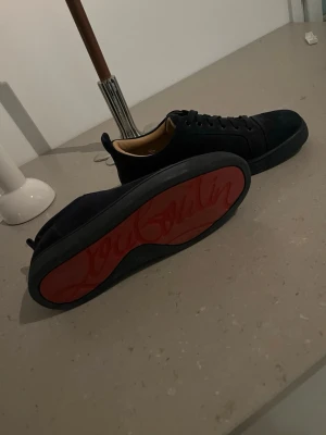 Svarta sneakers från Christian Louboutin - Snygga mörkblå sneakers i mocka från Christian Louboutin med klassisk röd sula och svarta skosnören. Låg modell med stilren design och beige innerfoder. Perfekta för dig som vill ha något exklusivt och trendigt. Köpta från Plick, Box  och dustbag ingår inte!