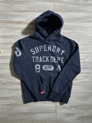 Mörkblå Superdry hoodie med tryck - Mörkblå hoodie från Superdry med stort vitt tryck på bröstet där det står 'Superdry Track Dept.' och siffrorna 84🤩 Ettiketen säger storlek S men skulle säga att den passar S-M🙌🏻 pris kan diskuteras