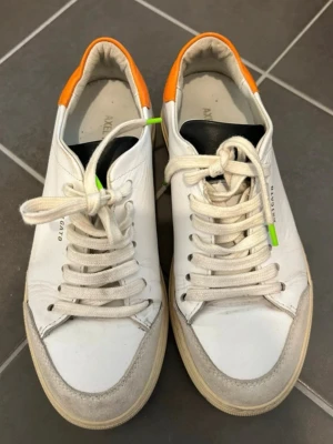 Vita sneakers från Axel Arigato - Snygga vita sneakers från Axel Arigato med orange detalj på hälen och grå mocka vid tån. Skorna har vita snören med små gröna detaljer och en klassisk låg siluett. Perfekta för dig som gillar stilrena och trendiga skor.