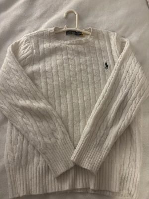 Vit kabelstickad tröja Polo Ralph Lauren - Kabelstickad vit tröja från Polo Ralph Lauren med klassisk rund halsringning och broderad logga på bröstet. Tröjan har ribbade muddar vid ärmslut och nederkant. Perfekt för dig som gillar tidlös och clean stil. qr Scan  finns