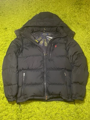 Svart dunjacka från Polo Ralph Lauren - Snygg svart dunjacka från Polo Ralph Lauren med huva och dragkedja framtill. Jackan har två framfickor med dragkedja och den klassiska röda loggan broderad på bröstet. Tillverkad i återvunnen polyester och dunfyllning för extra värme.