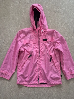 Vindjacka - Snygg rosa vindjacka med huva och dragkedja framtill. Jackan har två snedställda fickor med dragkedja och justerbara muddar vid ärmsluten. Tillverkad i lätt och tåligt material, perfekt för blåsiga dagar. Enkel och stilren design utan synligt märke. Finn fläckar.