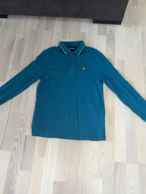 Blå långärmad skjorta Lyle & Scott - Snygg blå långärmad skjorta från Lyle & Scott med klassisk krage och knappar framtill. Har en liten gul logga på bröstet och randiga detaljer på kragen. Perfekt för dig som gillar stilrena plagg med en twist.