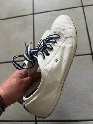 Vita sneakers från Tommy Hilfiger storlek 39 - Snygga vita sneakers från Tommy Hilfiger med klassiska marinblå och röda detaljer. Skorna har svarta snören med vita sömmar och är tillverkade i canvasmaterial. Hilfiger-loggan finns på sidan och bak på hälen. Perfekt för en stilren och avslappnad look.