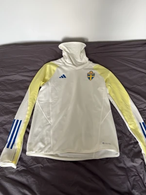 Svensk landslagströja Adidas vit - Vit långärmad landslagströja från Adidas med gula axelpartier och blå detaljer på ärmarna. Tröjan har hög krage och Sveriges fotbollsförbunds emblem på bröstet. Materialet är troligtvis polyester och har Climalite-teknologi för bra ventilation. Helt ny i storlek M inga konstigheter med den eller fläckar. Ny pris 1299kr 