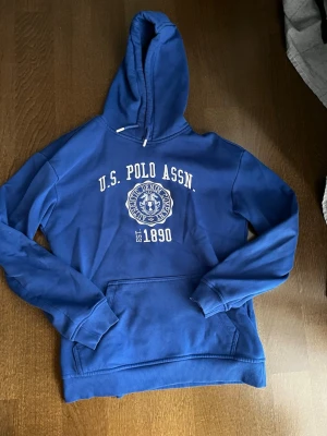 Blå hoodie från U.S. Polo Assn. - Snygg blå hoodie från U.S. Polo Assn. med vit logotyp och text på bröstet. Klassisk huva med dragsko och stor magficka framtill. Ribbstickade muddar vid ärmslut och nederkant. Perfekt för en avslappnad och cool look.