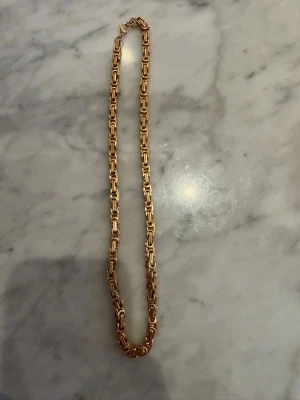 Guldfärgat kedjehalsband - Snyggt halsband i guldfärg med rejäla, rektangulära länkar och karbinlås. Halsbandet har en chunky känsla och passar perfekt för dig som gillar statement-smycken. Materialet ser ut som metall och designen är modern och stilren. Kedjan är i rostfritt stål och är fejkade 