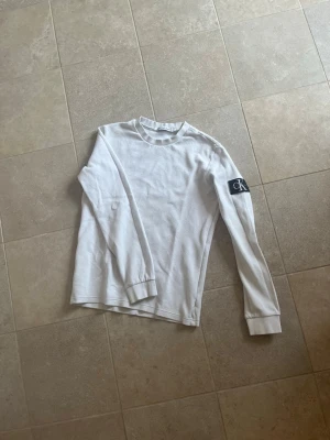 Vit långärmad tröja från Calvin Klein - Vit långärmad tröja från Calvin Klein med rund halsringning och ribbade muddar. Tröjan har en svart patch med CK-logga på ena ärmen och är tillverkad i ett mjukt, bekvämt material. Perfekt för en clean och stilren look.