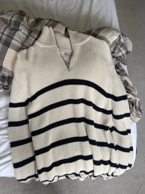 Randig stickad tröja från Zara - Snygg stickad tröja i offwhite med svarta horisontella ränder från Zara. Tröjan har halv dragkedja vid halsen, hög krage och ribbade muddar. Perfekt för en avslappnad och trendig look. 