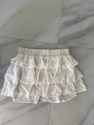 Vit volang kjol  - Supersöt vit kjol med flera lager volanger och elastisk midja. Perfekt för dig som gillar en lekfull och luftig stil. Materialet är lätt och känns skönt mot huden. 
