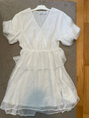 Vit kort klänning med volang - Söt vit kort klänning från NA-KD med puffärmar, v-ringning och volangdetaljer i midjan och nederkant. Klänningen har ett diskret blommigt mönster i tyget och är tillverkad i ett lätt, tunt material som passar perfekt till sommaren.