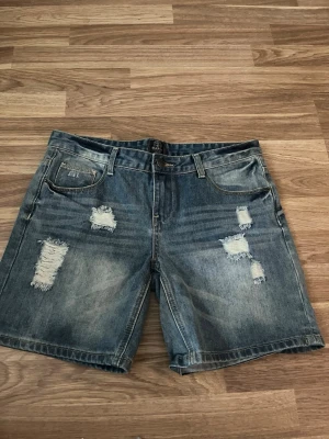 Blå denimshorts med slitningar - Snygga blå denimshorts från Second Legacy med flera slitna detaljer framtill och bakfickor. Klassisk femficksmodell med bälteshällor och knappgylf. Perfekta för en avslappnad stil under varma dagar.