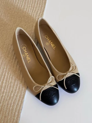 Beige och svarta ballerinaskor Chanel - Säljer ett par klassiska ballerinaskor från Chanel i beige läder med svart tåparti och diskret logga. Skorna har en liten rosett framtill och rundad tå. Perfekt för dig som gillar tidlös och elegant stil.