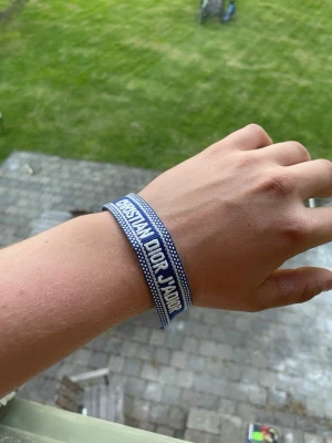 Blått Christian Dior J'Adior armband - Snyggt blått tygarmband från Christian Dior med texten 'CHRISTIAN DIOR J'ADIOR' i vitt. Armbandet har dekorativa tofsar i ändarna och ett diskret mönster längs kanterna. Perfekt accessoar för att lyfta din outfit med en exklusiv touch. Tveka inte å fråga eller pruta