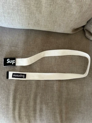 Vitt Supreme bälte med svart spänne - Säljer ett vitt bälte från Supreme med svart metallspänne där det står 'Sup' i vitt. Bältet har även en Supreme-logga på tygdelen. Perfekt accessoar för att lyfta din outfit och visa din streetwear-stil.
