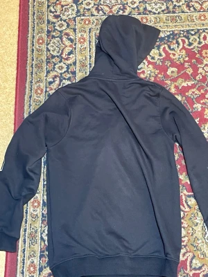Svart hoodie från Lacoste - Snygg svart hoodie från Lacoste med stor vit text på bröstet och ärmen. Klassisk huva med dragsko och ribbade muddar. Ikoniska Lacoste-loggan med krokodil på bröstet. Perfekt för en avslappnad och trendig look.