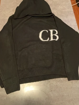 Svart hoodie med CB-tryck - Säljer en svart hoodie med stor huva och vita bokstäver 'CB' på bröstet. Hoodien har ribbade muddar vid ärmslut och nederkant. Materialet ser ut att vara mjukt och stickat, perfekt för en avslappnad stil.