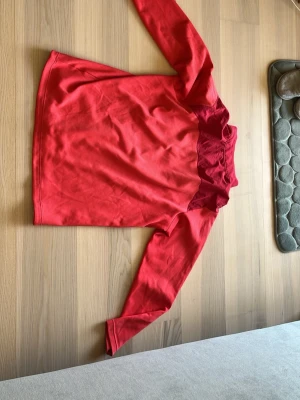 Röd långärmad träningströja Nike - Snygg röd långärmad golf träningströja från Nike med halv dragkedja och hög krage. Tröjan har två olika röda nyanser och är tillverkad i ett mjukt, funktionsmaterial som passar perfekt till träning eller uppvärmning. Storlek xl men passar L
