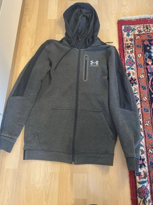 Mörkgrå tracksuit från Under Armour - Mörkgrå hoodie från Under Armour med dragkedja, fickor och logga på bröstet. Tillverkad i mjukt och stretchigt material, har en stor huva och detaljer i svart på ärmarna. Perfekt för dig som gillar sportig och clean stil.