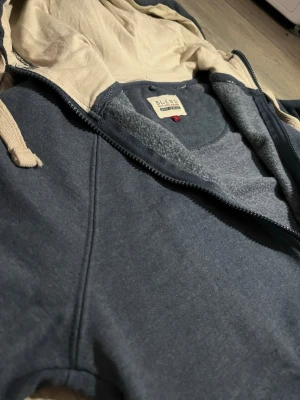 Mörkblå zip hoodie från Blend! - Snygg mörkblå hoodie från Blend med dragkedja framtill och beige insida i huvan. Hoodien har dragsko i huvan och ribbade muddar vid ärmslut och nederkant. Perfekt för en avslappnad stil och enkel att matcha med jeans eller joggers. Den har ett skönt material innuti tröjan! 