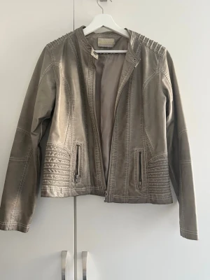 Grå skinnjacka från MarcCain - Snygg grå skinnjacka från MarcCain med coola detaljer på axlar och nedtill. Jackan har dragkedja framtill, två fickor med dragkedja och en stilren, rak passform. Perfekt för dig som vill ha en tidlös och trendig look. Jackan köpt secondhand och är i storlek XL men passar bra för mig som är M. 