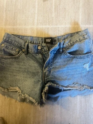 Blå jeansshorts från 157 med fransar - Säljer ett par klassiska blå jeansshorts från 157 med råa, fransiga benslut och slitna detaljer framtill. Shortsen har fem fickor, knappgylf och normal passform. Perfekta till sommaren och lätta att matcha med olika stilar.