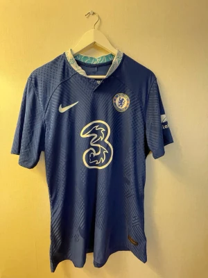 Chelsea matchtröja Nike blå T.Silva - Chelsea FC matchtröja i blått med vita detaljer, tillverkad av Nike. Tröjan har klubbmärke, Nike-logga och sponsortryck på framsidan samt T.SILVA och nummer 6 på ryggen. Tillverkad i lätt och ventilerande material, perfekt för fotbollsträning eller match.