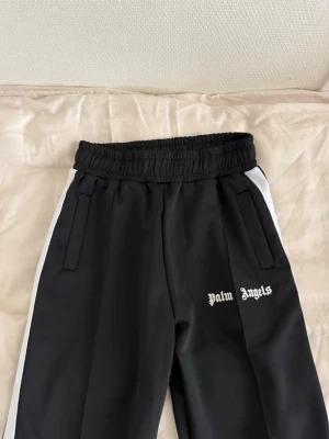 Palm Angels svarta mjukisbyxor storlek M - Palm Angels mjukisbyxor i svart med vita sidoränder och logga framtill. Byxorna har resår i midjan och två sidofickor. Snygg streetwear-look med avslappnad passform och klassisk design. Perfekta för dig som gillar trendiga och bekväma plagg.