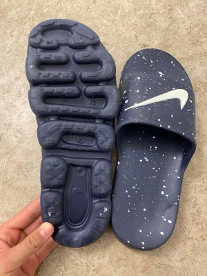 Blå Nike tofflor med vita stänk - Säljer ett par blåa Nike tofflor med vita färgstänk och stor vit Swoosh-logga på ovandelen. Sulan har unik struktur och mönster för extra grepp. Tillverkade i syntetmaterial, perfekta för chill dagar hemma eller efter träningen.