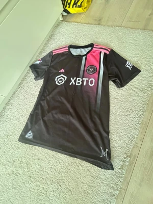Inter Miami CF matchtröja svart/rosa - Snygg Inter Miami CF matchtröja från Adidas i svart med rosa och grå detaljer. Tröjan har klubbmärke på bröstet, sponsorlogga och klassiska Adidas-ränder på axlarna. Perfekt för fotbollsträning eller att bära som supporter.