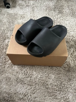Svarta Yeezy Slide tofflor - Säljer ett par svarta Yeezy Slide tofflor med öppen tå och chunky sula. Tillverkade i mjukt och flexibelt syntetmaterial för maximal komfort. Minimalistisk design utan synliga loggor, perfekt för en clean och modern look.
