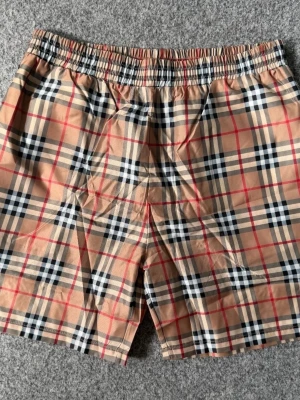 Beige rutiga shorts Burberry-style - Säljer ett par klassiska beige shorts med svart, vit och röd rutig design i Burberry-stil. Shortsen har elastisk midja och är tillverkade i ett lätt bomullsmaterial, perfekta för sommaren. Passar dig som gillar tidlös och trendig look.