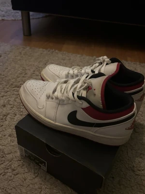 Air Jordan 1 Low White/Red/Black - Säljer ett par Air Jordan 1 Low sneakers i vitt läder med svarta och röda detaljer. Endast använd någon fåtal gång. Har liten defekt på skosnöret men man kan billigt byta eller tvätta bort det. Skorna går på farfetch för lite över 4000kr.