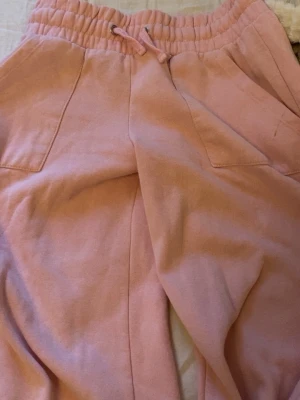 Ljusa rosa mjukisbyxor med fickor - Säljer ett par ljusa rosa mjukisbyxor med elastisk midja och dragsko. Byxorna har två stora fickor framtill och är gjorda i ett mjukt bomullsmaterial. Perfekta för chill dagar och har en relaxed fit som är riktigt skön.