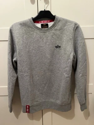 Grå sweatshirt från Alpha Industries - Snygg grå sweatshirt från Alpha Industries med rund halsringning och diskret logga på bröstet. Tröjan har ribbade muddar vid ärmslut och nederkant samt en röd detalj vid fållen. Perfekt till ett avslappnat streetwear-look.