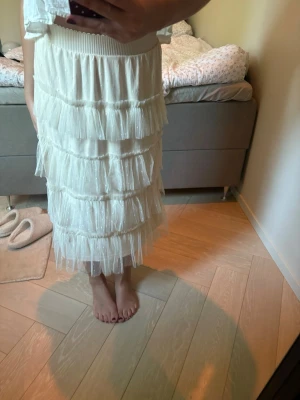Vit volangkjol med mesh - Säljer en superfin vit långkjol med flera lager volanger i mesh. Kjolen har resår i midjan och ett skirt, lätt material som ger en luftig känsla. Perfekt för dig som gillar romantisk och trendig stil.