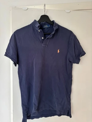 Marinblå pikétröja från Ralph Lauren - Klassisk marinblå pikétröja från Ralph Lauren med korta ärmar och den ikoniska broderade loggan i orange på bröstet! Perfekt för en avslappnad men ändå stilren look. Den är missfärgad men fortfarande fin att använda! Krympt från M till XS/S