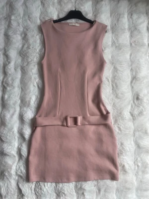 Rosa kort klänning med bälte - Snygg rosa kort klänning i figurnära modell med rund halsringning och matchande bälte i midjan. Ärmlös design och enkel, stilren look som passar perfekt till många tillfällen. Materialet känns mjukt och följsamt. Storlek S.